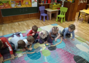 Grupka dzieci na dywanie. Ułożone drewniane tory. Obserwują jak pociąg porusza się po torach.